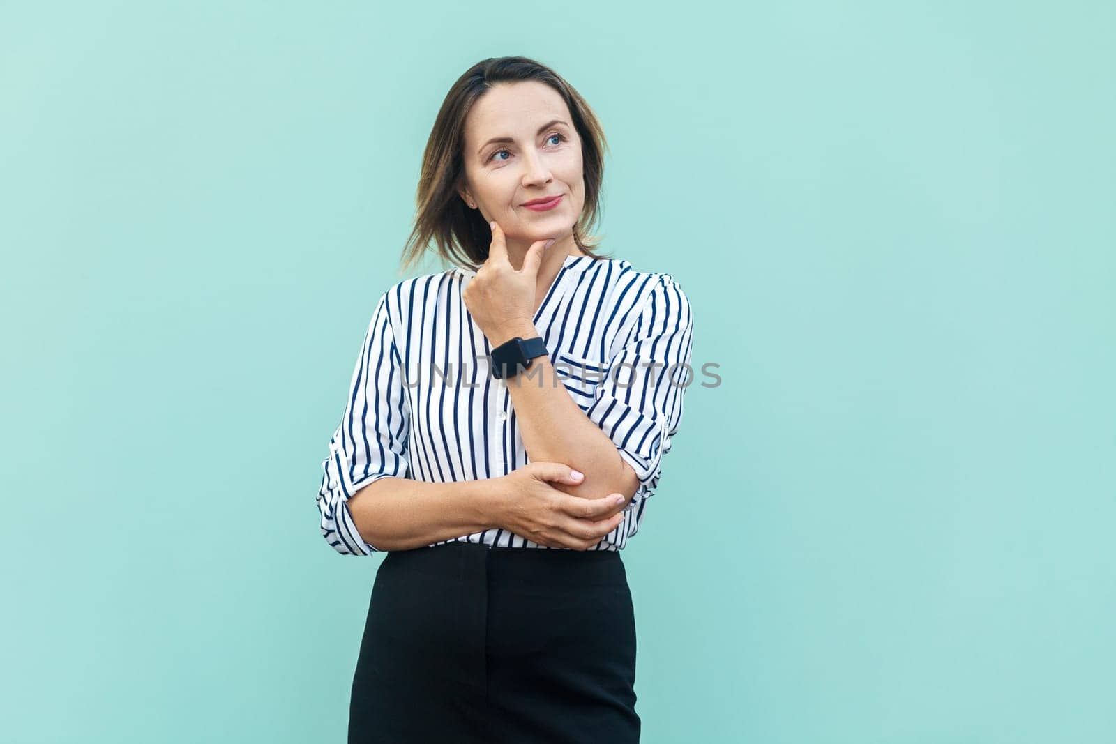 Portrait of pensive attractive middle aged woman wearing striped shirt standing holding chin, thinking about future plans. Indoor studio shot isolated on light blue background.