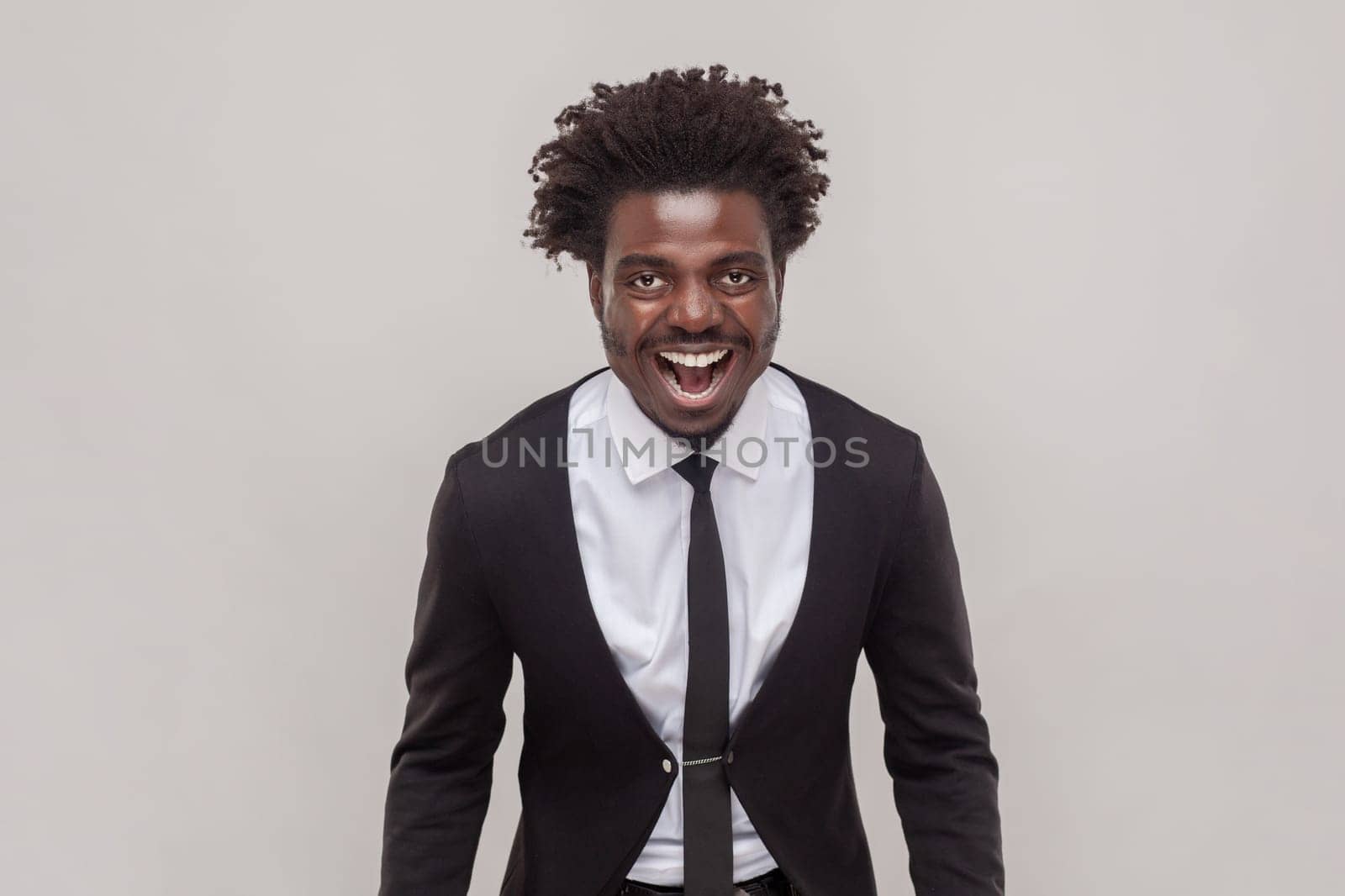 Man with petit goatee shouting loudly having aggression, screaming with wide opened mouth, having unhappy expression, wearing white shirt and tuxedo. Indoor studio shot isolated on gray background.