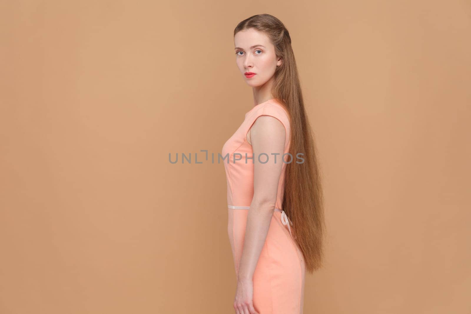 Side view portrait of serious strict woman with long hair looking at camera with bossy expression, being in bad mood, wearing elegant dress. Indoor studio shot isolated on brown background.