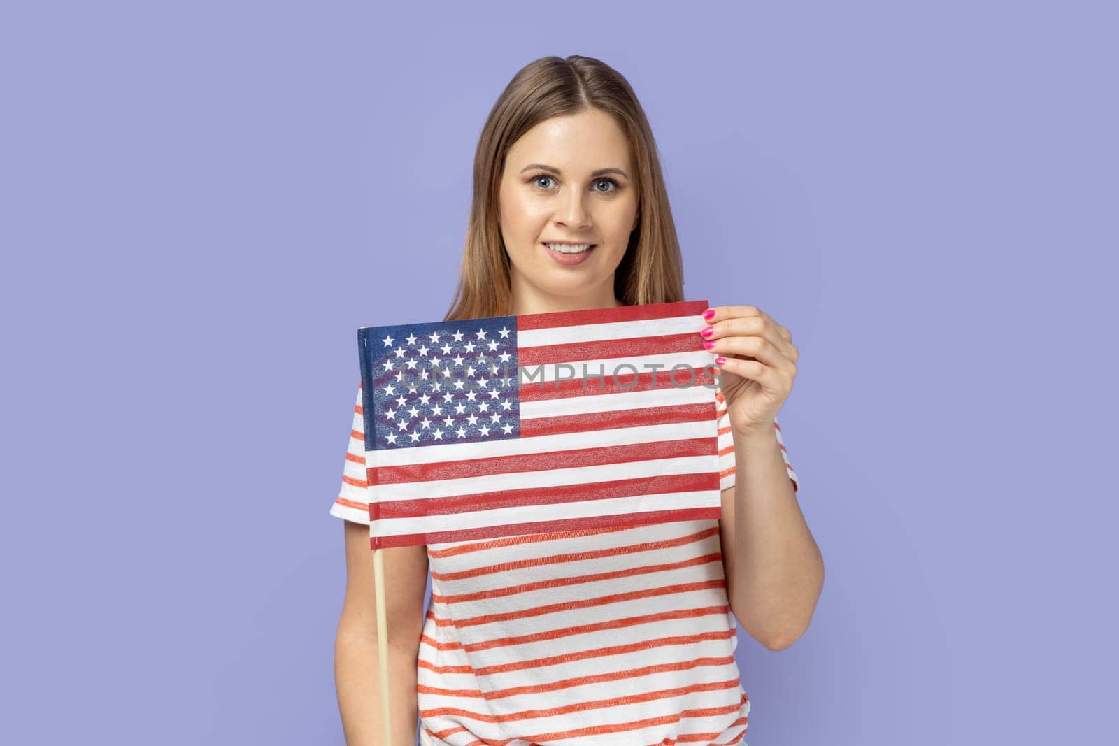 Portrait of blond woman wearing T-shirt holding in hand flag of united states of america, celebrating independence day, having calm facial expression. Indoor studio shot isolated on purple background.