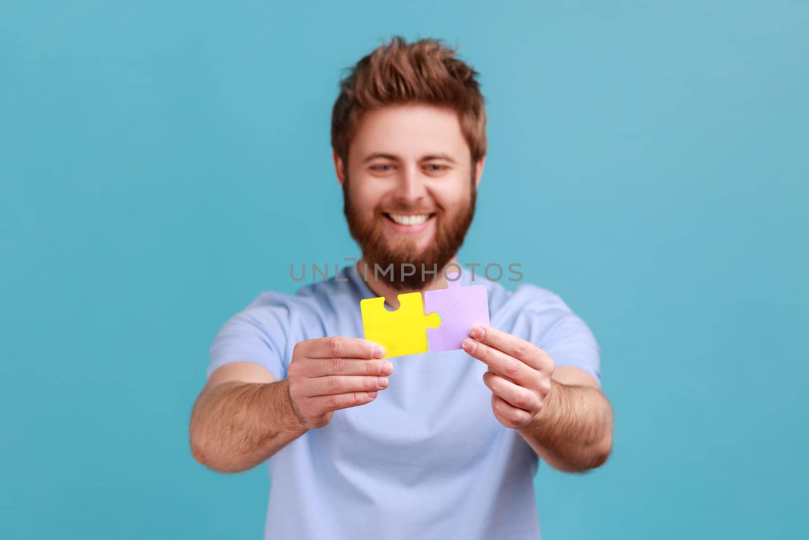 Portrait of positive happy bearded man combining to different colorful pieces of puzzle, making right solution, creating business association. Indoor studio shot isolated on blue background.