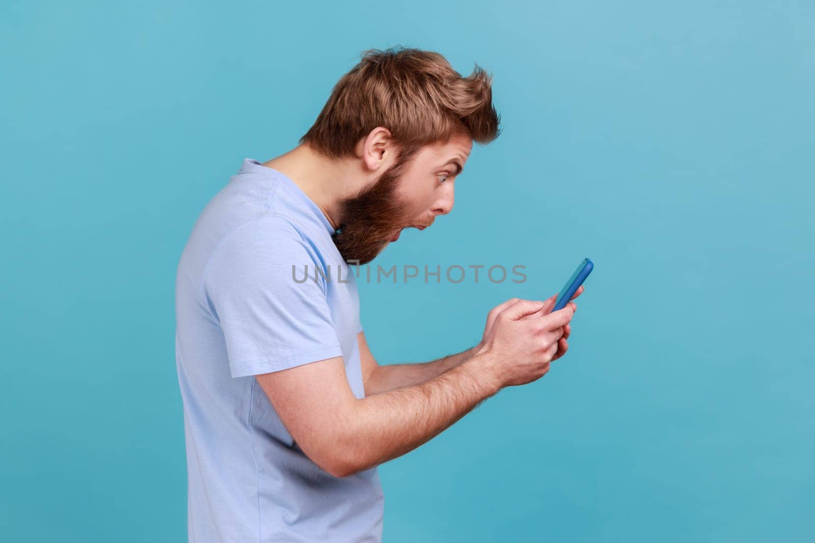 Side view of astonished surprised young adult bearded man standing, using smartphone and watching video with amazed face and open mouth. Indoor studio shot isolated on blue background.