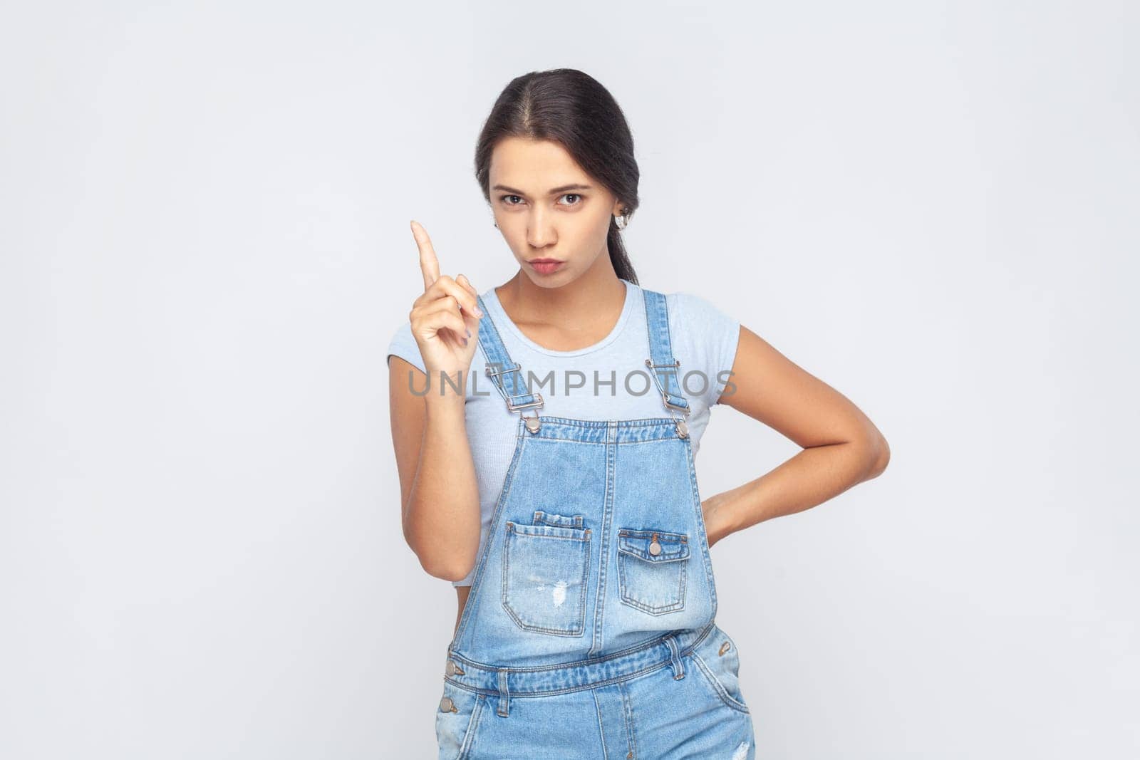 Portrait of serious strict bossy woman wearing denim overalls standing with raised index finger, warning you, looking seriously at camera. Indoor studio shot isolated on gray background.