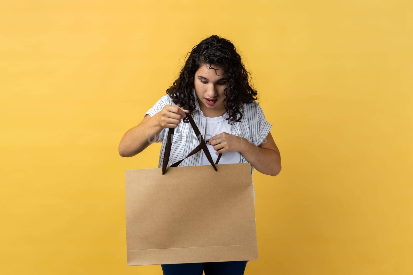 Portrait of amazed attractive young adult woman with dark wavy hair holding and looking inside paper shopping bag, being shocked by content. Indoor studio shot isolated on yellow background.