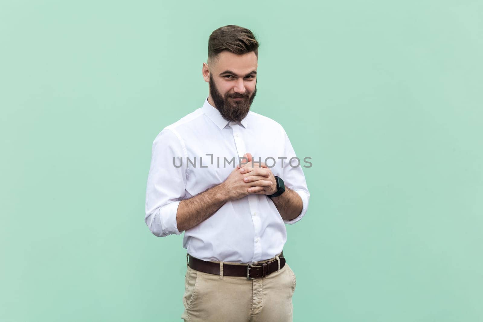 Portrait of young adult bearded businessman wearing white shirt looking at camera with cunning tricky face and smirk, planning evil trick. Indoor studio shot isolated on light green background.