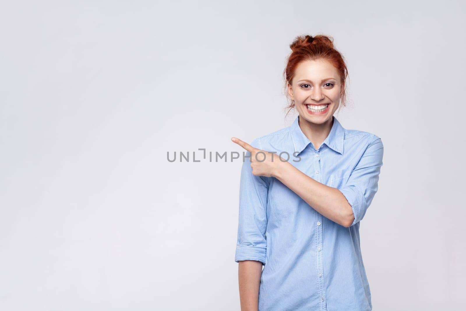 Portrait of ginger woman wearing blue shirt looking at camera and pointing away, presenting copy space for advertisement or promotion. Indoor studio shot isolated on gray background.
