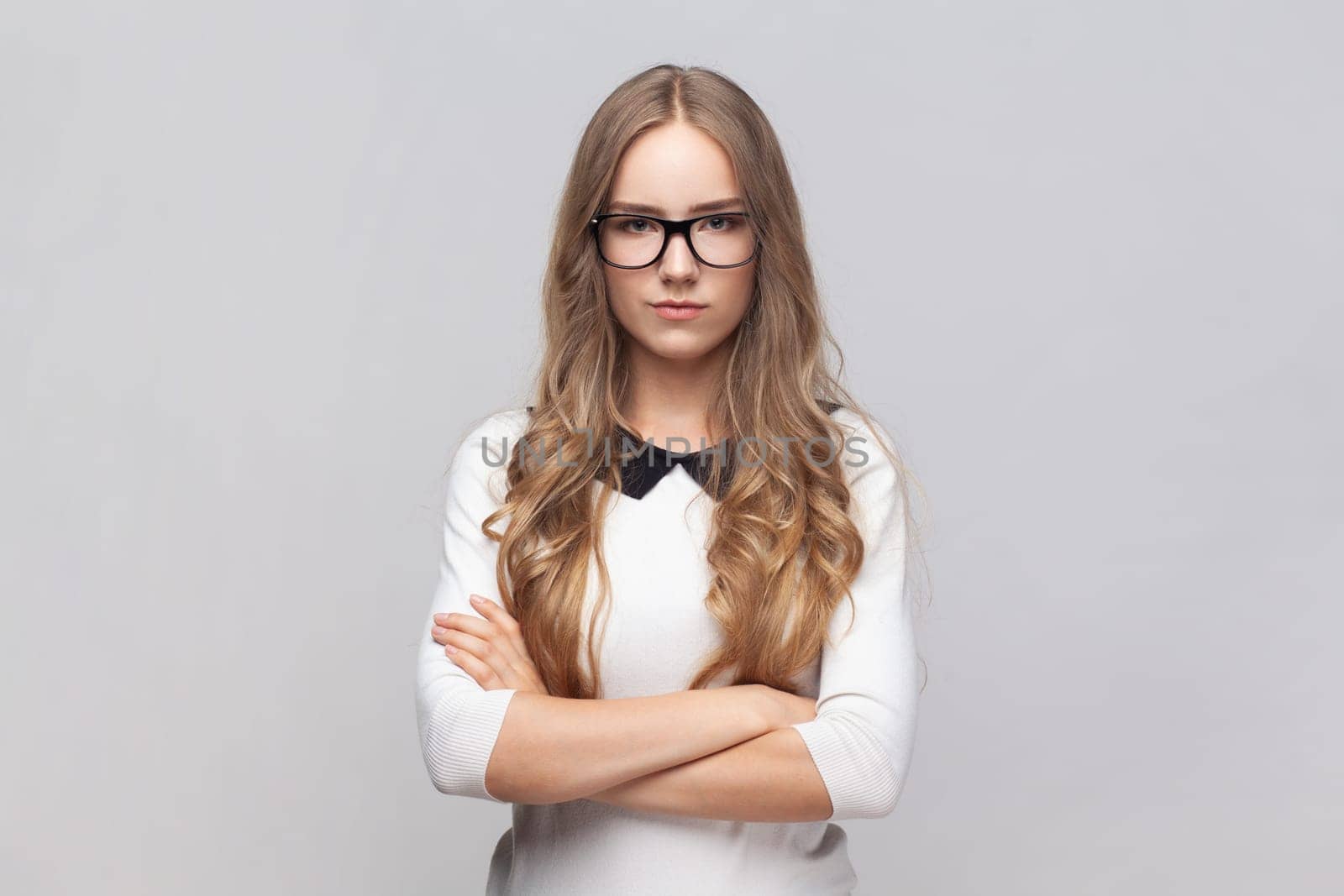 Portrait of serious strict young adult woman in glasses with wavy blond hair standing looking at camera with bossy expression, keeps hands crossed. Indoor studio shot isolated on gray background.