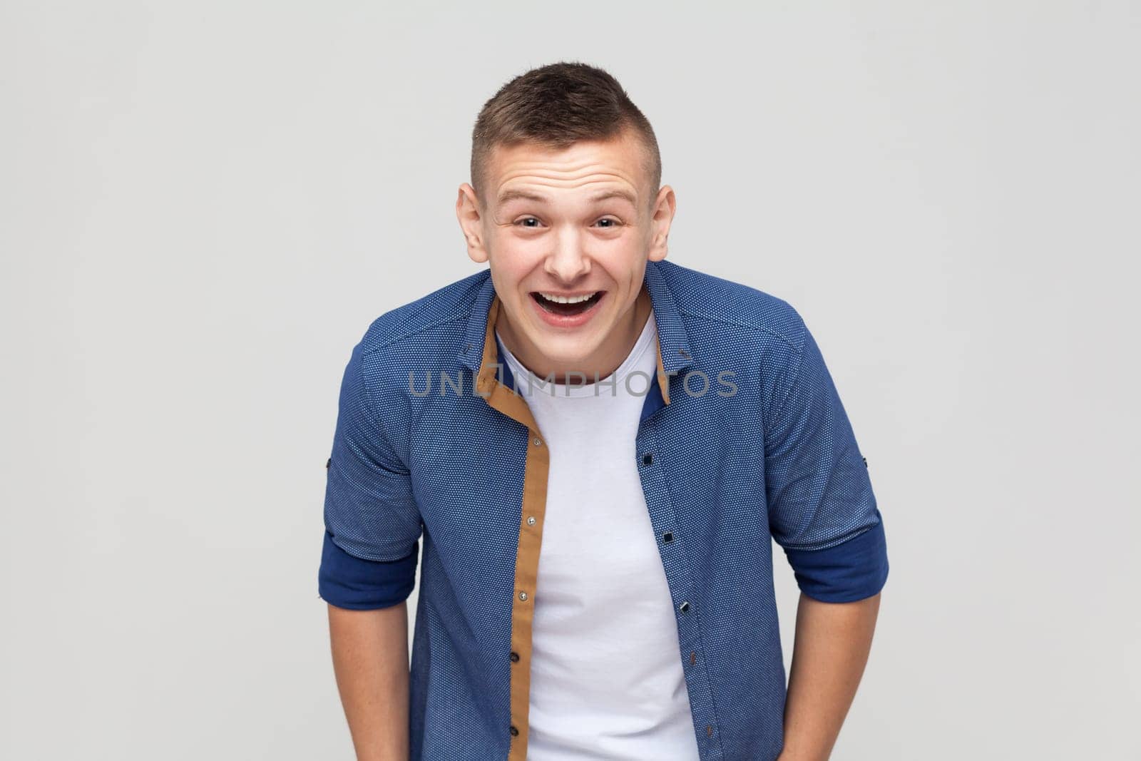 Portrait of overjoyed extremely happy handsome teenager boy wearing blue shirt hunching from laughter, laughing out loud, crazy face. Indoor studio shot isolated on gray background.