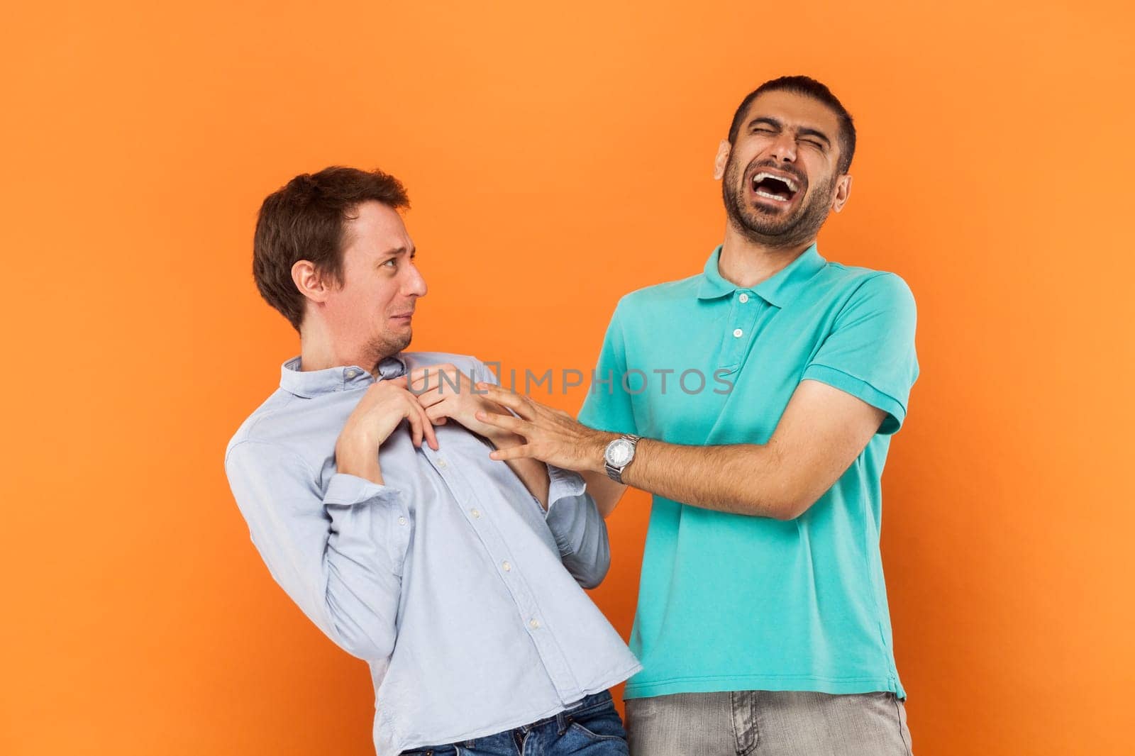Portrait of two handsome young adult men posing together, friend mocking and laughs at another guy, laughing lout out with closed eyes. Indoor studio shot isolated on orange background.
