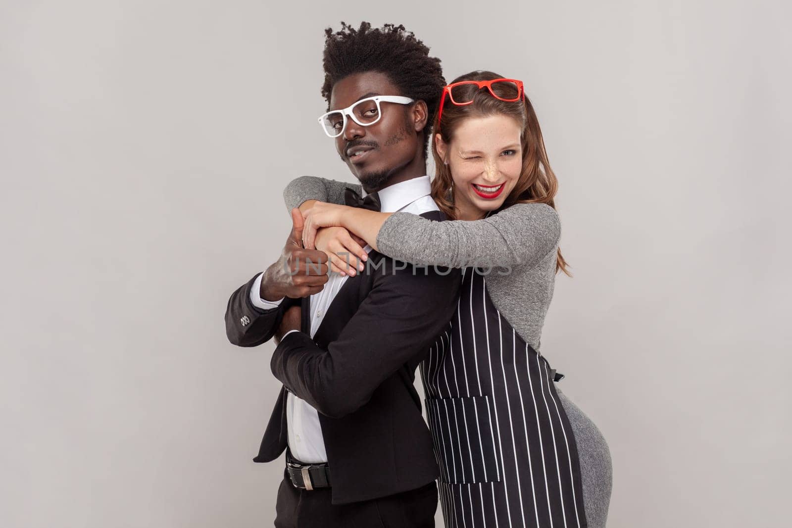 Portrait of young adult couple woman and man in white glasses standing together, guy showing thumb up, female winking playfully to camera. Indoor studio shot isolated on gray background.