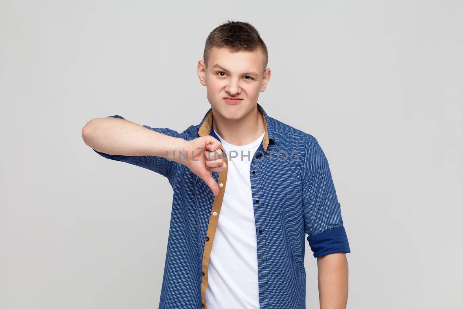 Portrait of naughty dissatisfied teenager boy wearing blue shirt frowning angrily and showing thumbs down gesture, expressing disapproval, dislike. Indoor studio shot isolated on gray background.