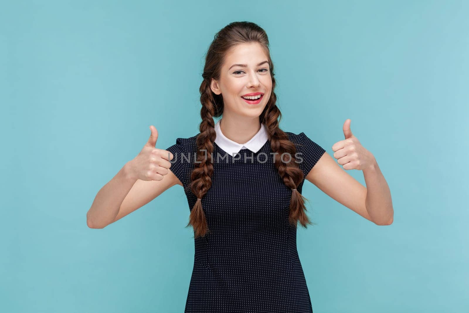 Portrait of attractive positive smiling woman with braids keeps thumb raised, being in good mood, shows her agreement, wearing black dress. woman Indoor studio shot isolated on blue background.