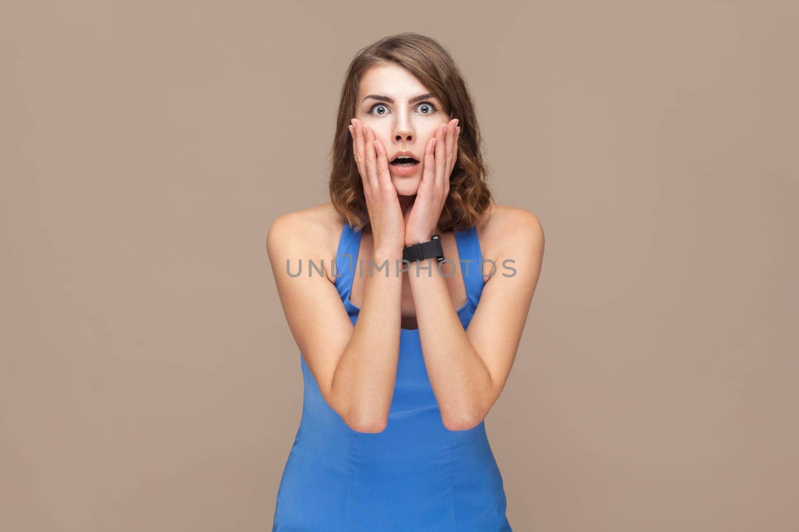 Portrait of shocked surprised beautiful woman with wavy hair standing with palms on her cheeks, sees something astonished, wearing blue dress. Indoor studio shot isolated on light brown background.