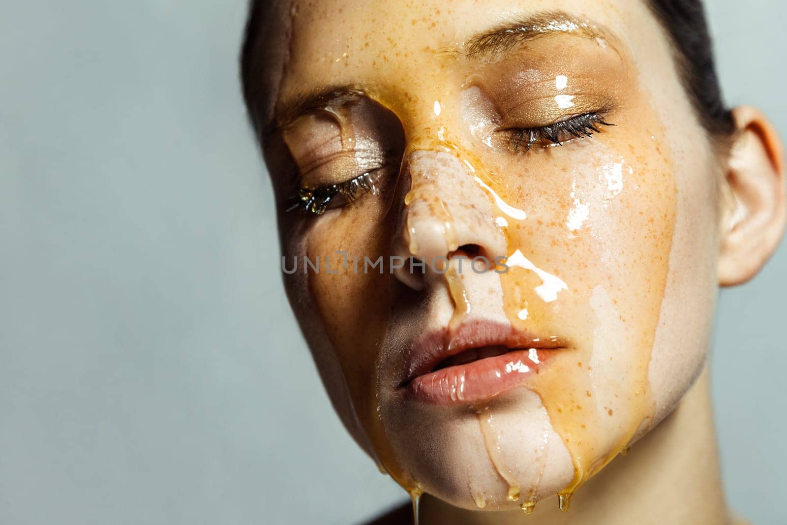 Closeup portrait of attractive calm young woman with freckles and honey on face, keeps eyes closed, having serious expression. Indoor studio shot isolated on gray background.