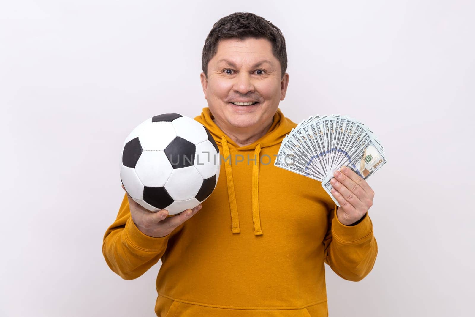 Smiling middle aged man showing soccer ball and fun of hundred dollar bills, winning lot of money betting for sport, wearing urban style hoodie. Indoor studio shot isolated on white background.