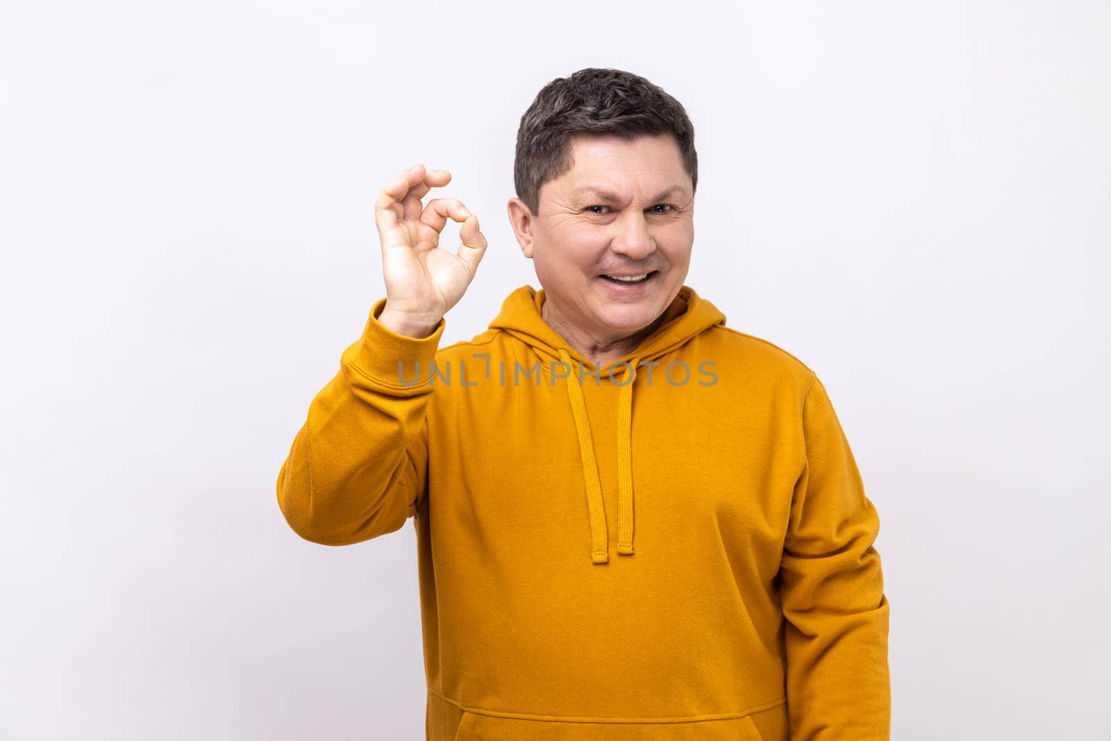 Portrait of playful optimistic handsome brunette man showing okay finger gesture and looking at camera, wearing urban style hoodie. Indoor studio shot isolated on white background.