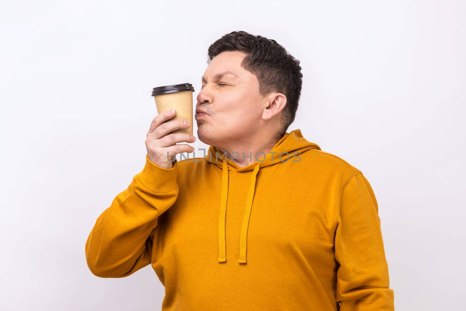 Portrait of joyful middle with dark hair kissing disposable cup of take away coffee, enjoying beverage, wearing urban style hoodie. Indoor studio shot isolated on white background.