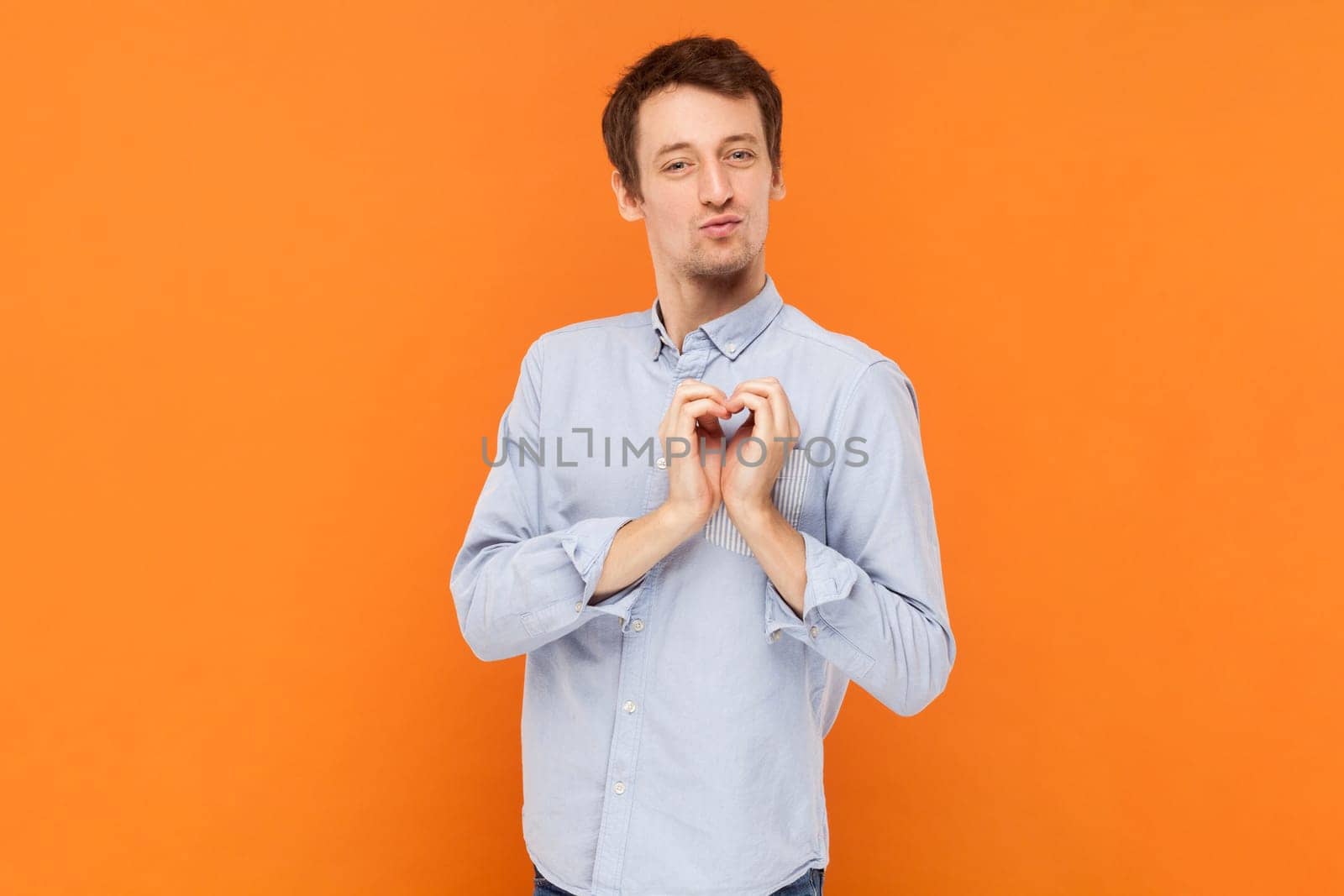 Portrait of funny romantic handsome man standing and showing heart shape gesture, falling in love, wearing light blue shirt. Indoor studio shot isolated on orange background.