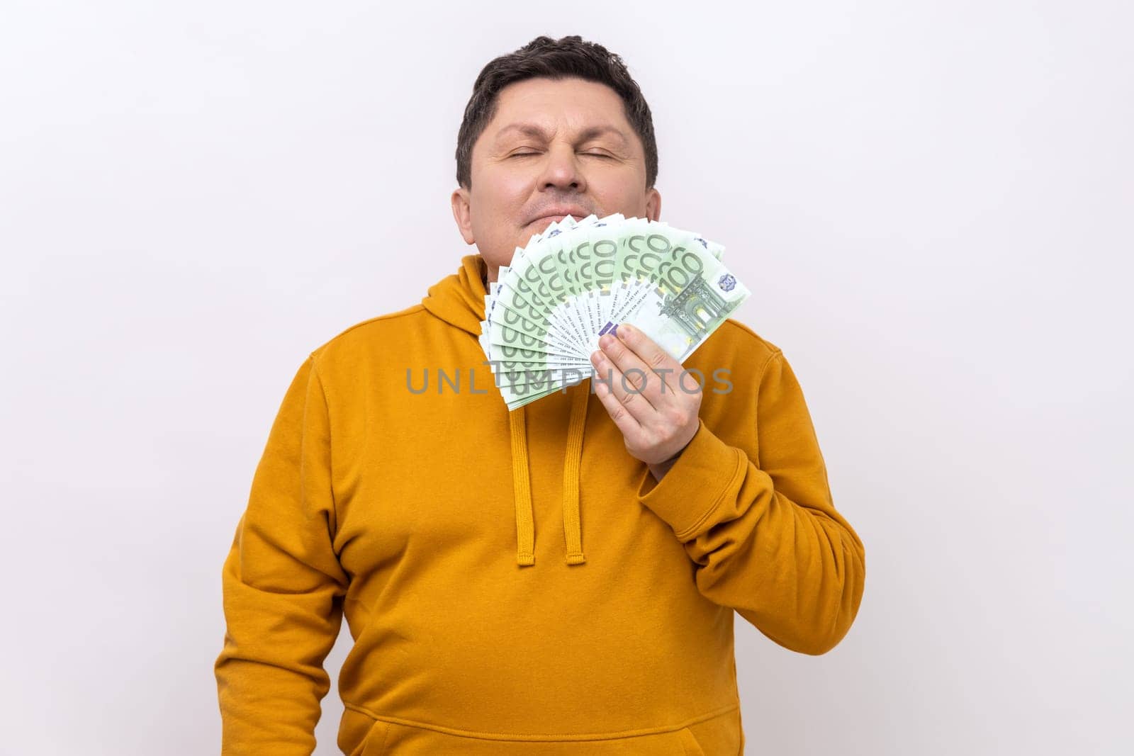 Greedy mercantile man smelling fan of cash, enjoying aroma of hundred euro bills in his hand, satisfied with big profit, wearing urban style hoodie. Indoor studio shot isolated on white background.