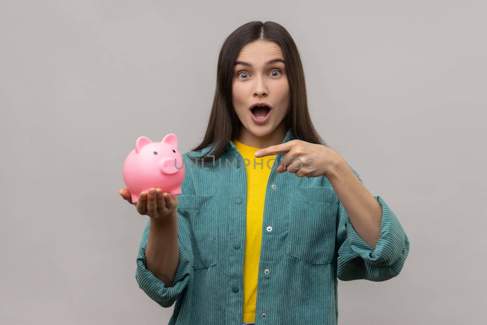 Portrait of amazed woman with dark hair standing pointing at piggy bank in her hand, advantageous bank offer, wearing casual style jacket. Indoor studio shot isolated on gray background.