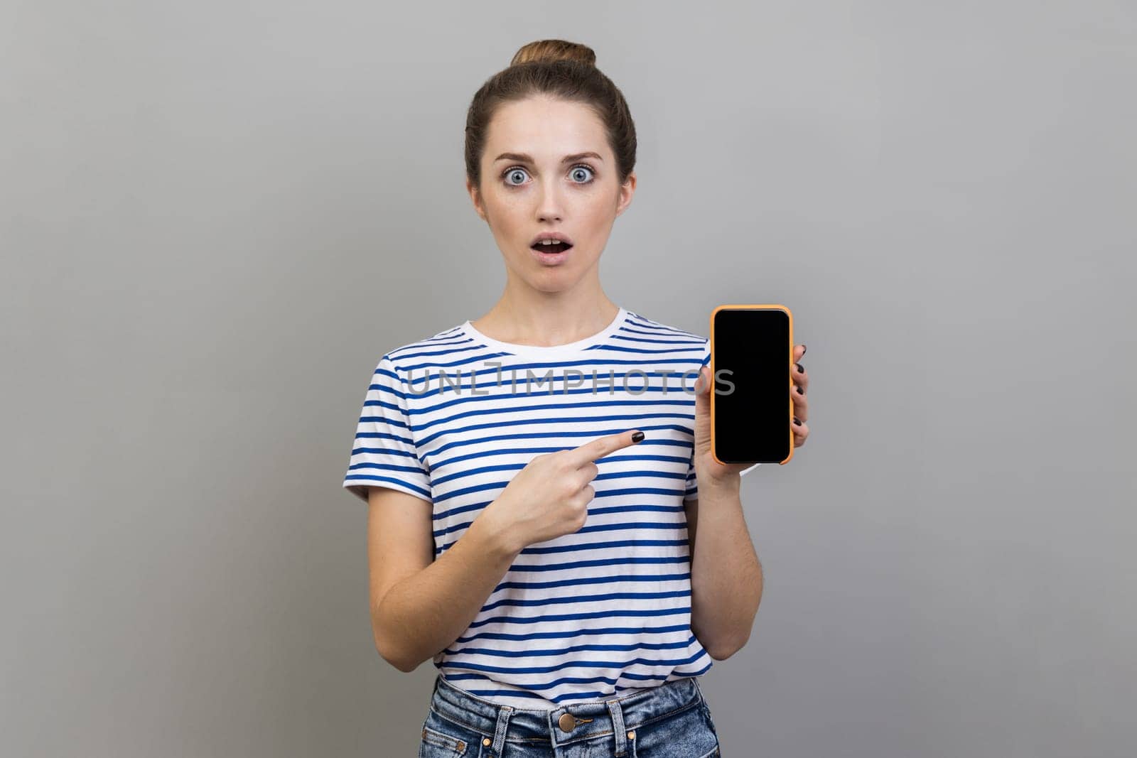 New mobile app. Portrait of woman wearing striped T-shirt pointing at cell phone and looking with surprised expression, shocked by mobile tariffs. Indoor studio shot isolated on gray background.