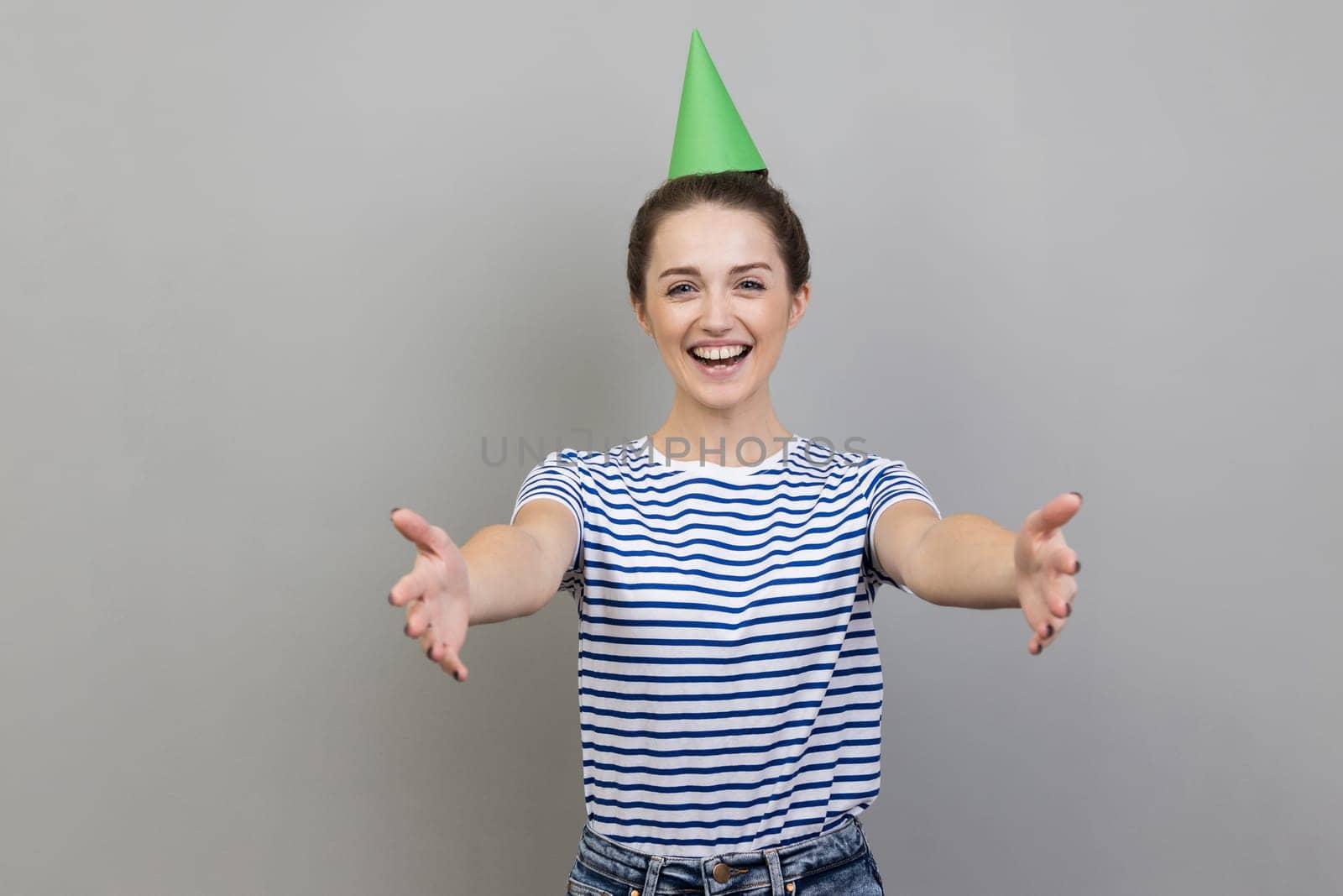 Pleased smiling woman wearing striped T-shirt and party cone, inviting her friends to birthday party by Khosro1