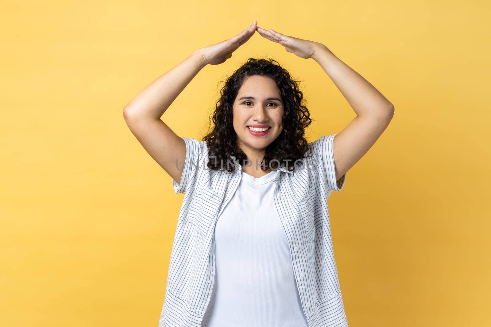 Portrait of woman with dark wavy hair and charming smile holding hands in gesture of house roof over head and smiling, feeling safe. Indoor studio shot isolated on yellow background.