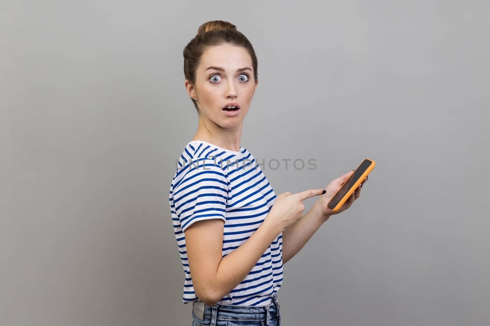 Portrait of amazed astonished woman blogger wearing striped T-shirt using smart phone, looking at camera with open mouth, sees shocking content. Indoor studio shot isolated on gray background.