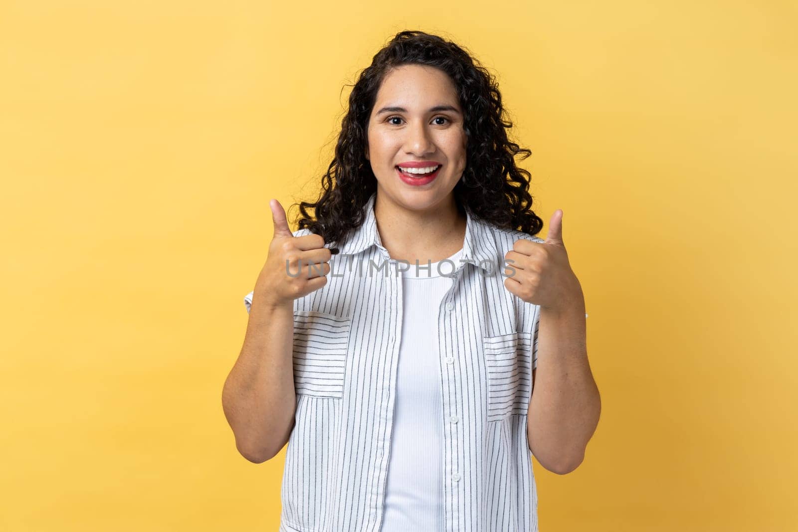 Portrait of woman with dark wavy hair looking at camera with toothy smile and showing thumbs up, approval sign, satisfied with service, good feedback. Indoor studio shot isolated on yellow background.