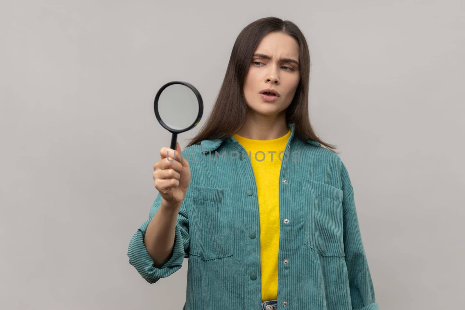 Curious woman with dark hair looking away through magnifying glass, spying, finding out something, inspecting, wearing casual style jacket. Indoor studio shot isolated on gray background.