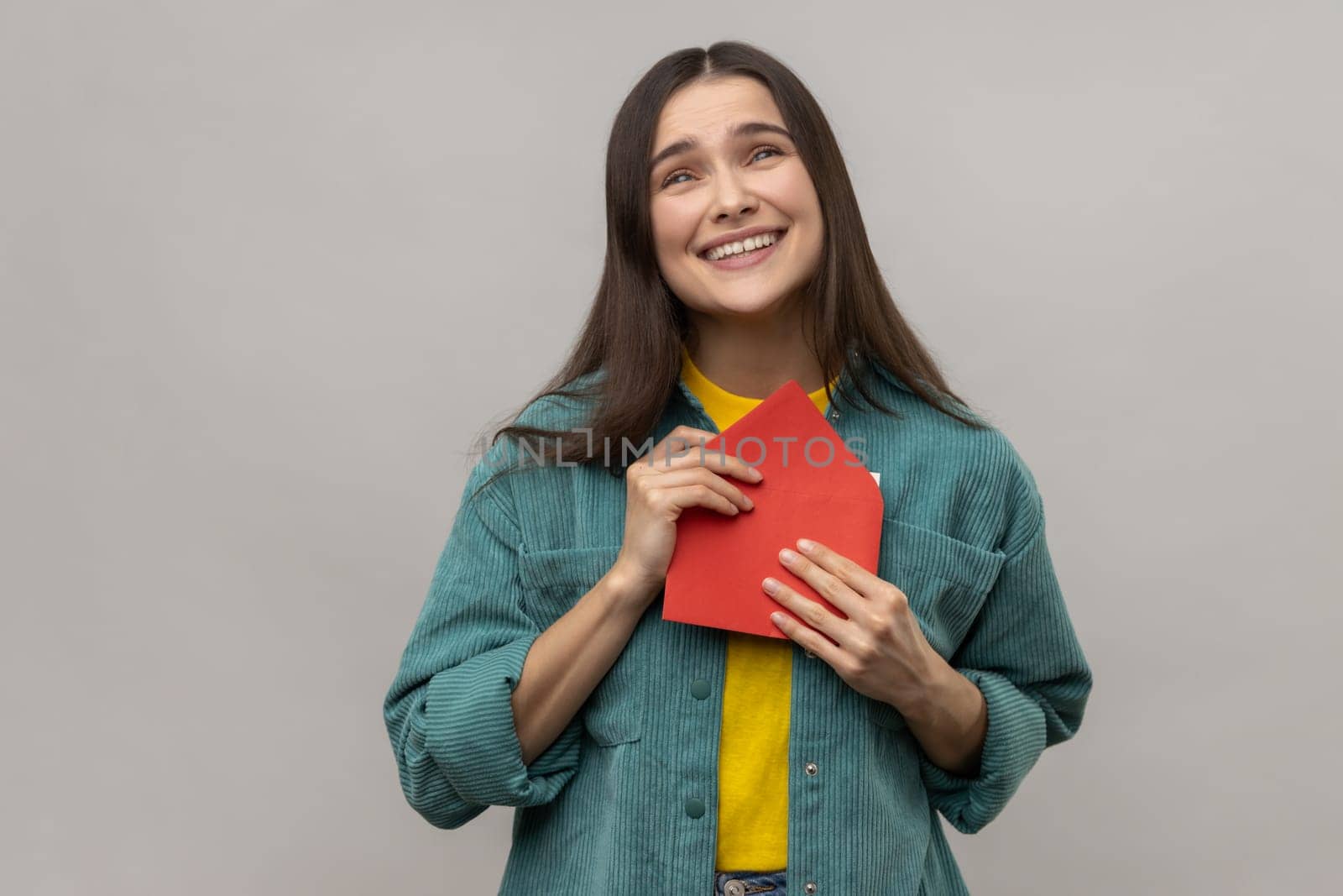 Portrait of pleased young woman holding envelope and reading letter, with happy expression, being touched, wearing casual style jacket. Indoor studio shot isolated on gray background.