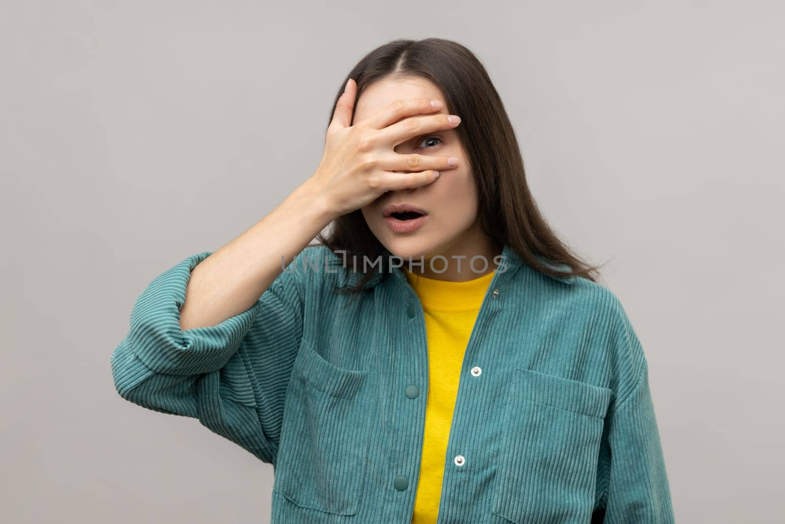 Portrait of curious dark haired woman spying, hiding and peeping through fingers, looking for secrets, rumors, wearing casual style jacket. Indoor studio shot isolated on gray background.