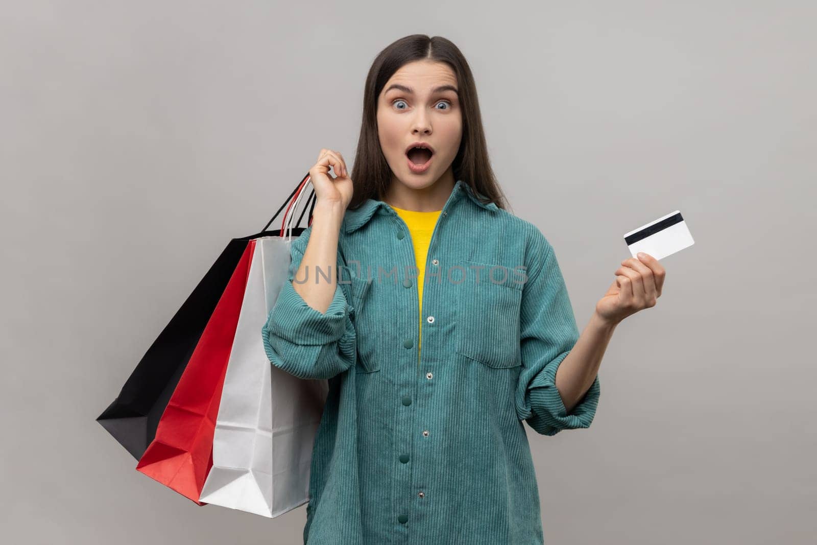 Surprised woman holding and showing limitless credit card and shopping bags, shocked with prices, final sale, wearing casual style jacket. Indoor studio shot isolated on gray background.