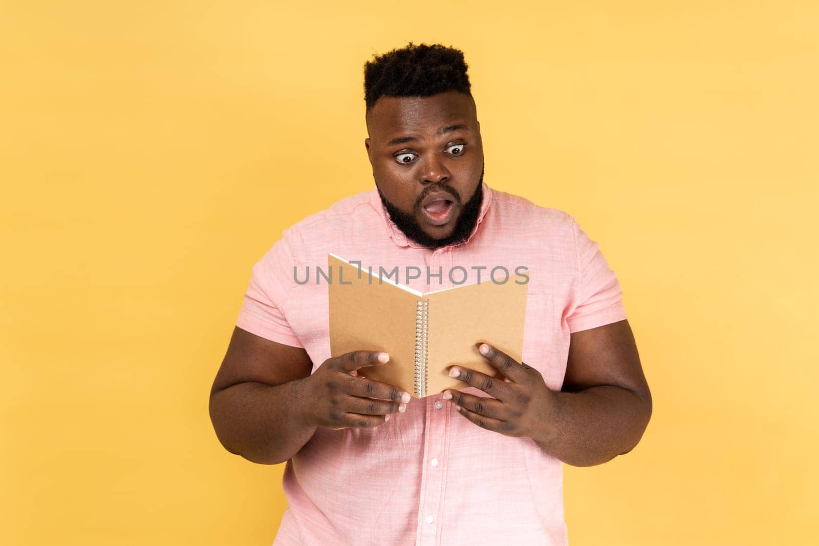 Shocking facts. Astonished man wearing pink shirt reading book with surprised expression, amazed by story, being shocked by interesting information. Indoor studio shot isolated on yellow background.