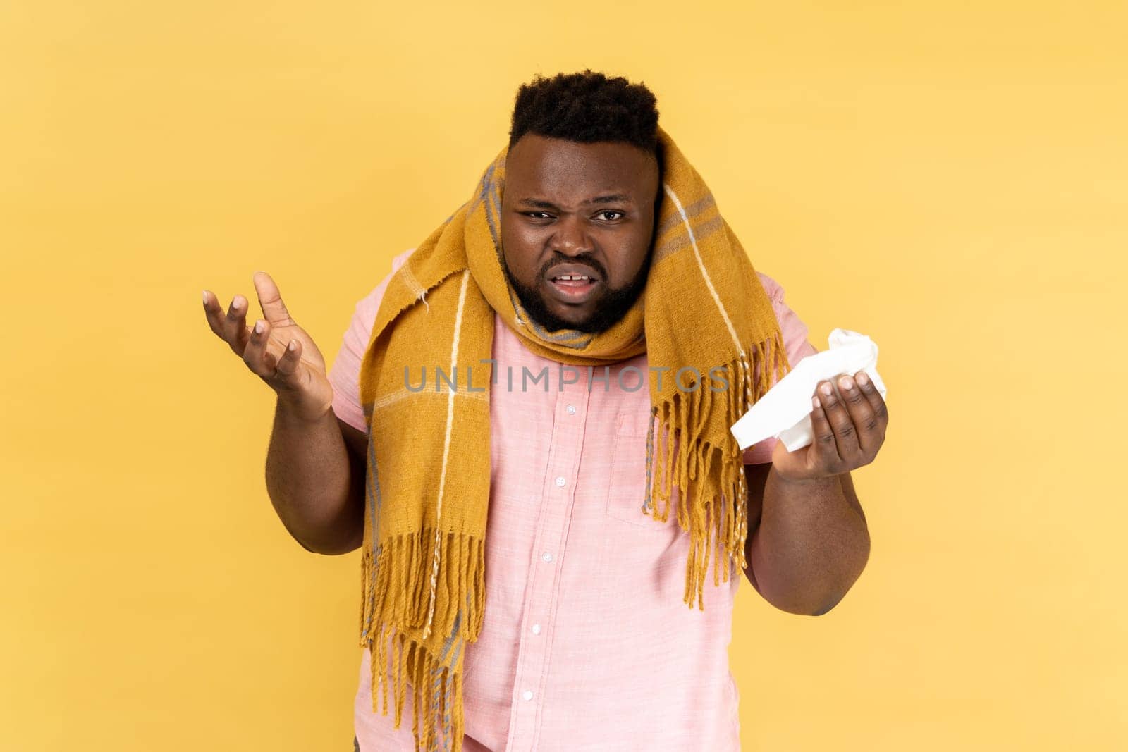 Seasonal influenza. Portrait of man wearing pink shirt wrapped in scarf looking with displeased grimace, treating flu, cough and fever. Indoor studio shot isolated on yellow background.