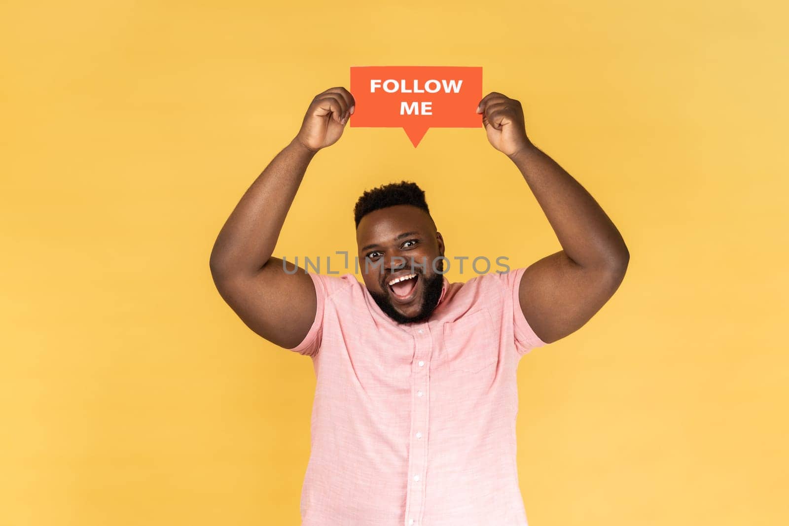 Portrait of cheerful excited bearded young adult man blogger wearing pink shirt standing card with fallow me inscription. Indoor studio shot isolated on yellow background.