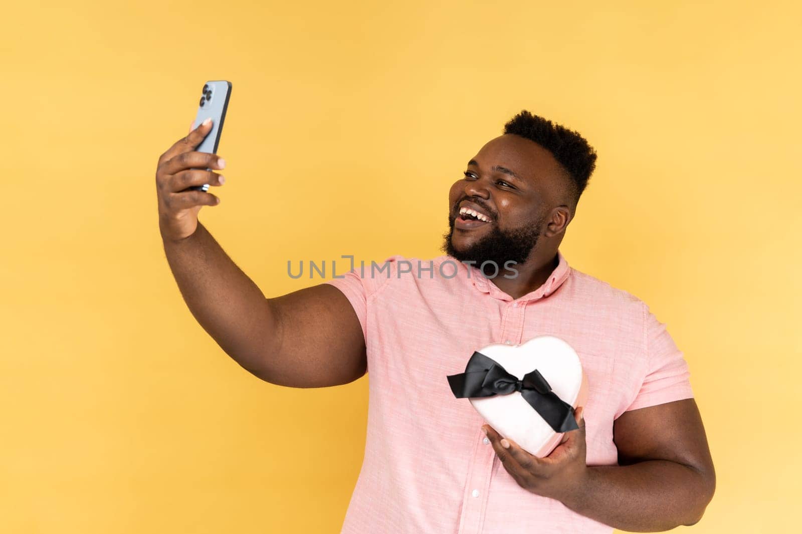 Portrait of smiling joyful man wearing pink shirt holding heart shaped gift box and making selfie, video call or broadcasting livestream. Indoor studio shot isolated on yellow background.