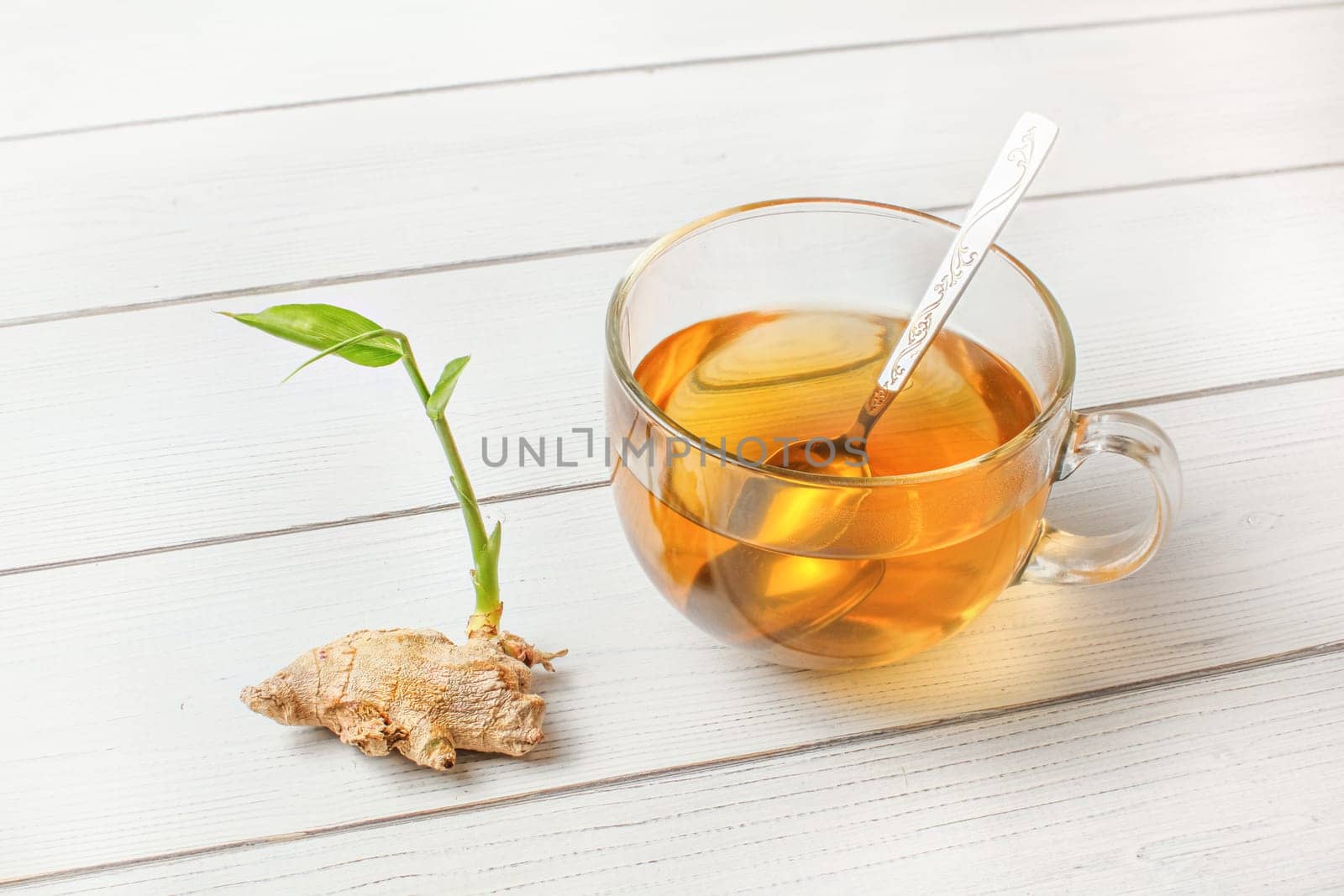Glass cup of freshly brewed amber tea, dry ginger root with green sprout next to it on white boards desk.