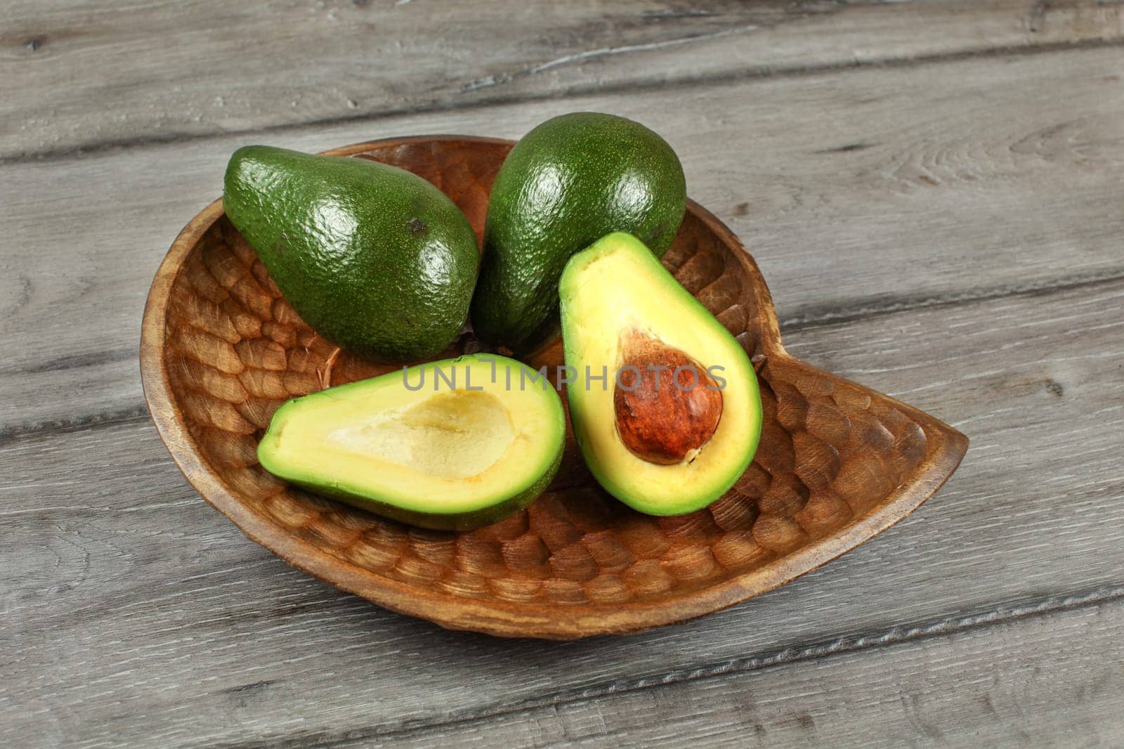 Three avocados, one of them cut to half, seed visible, in carved wooden bowl.