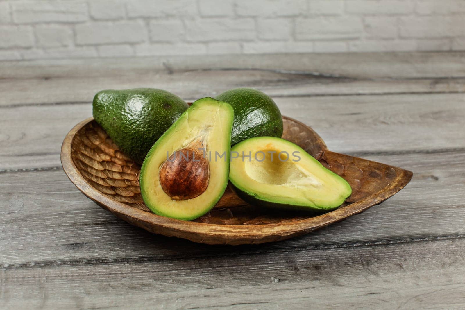 Avocado cut in half, seed and pulp visible, two more whole pears on carved wooden bowl.
