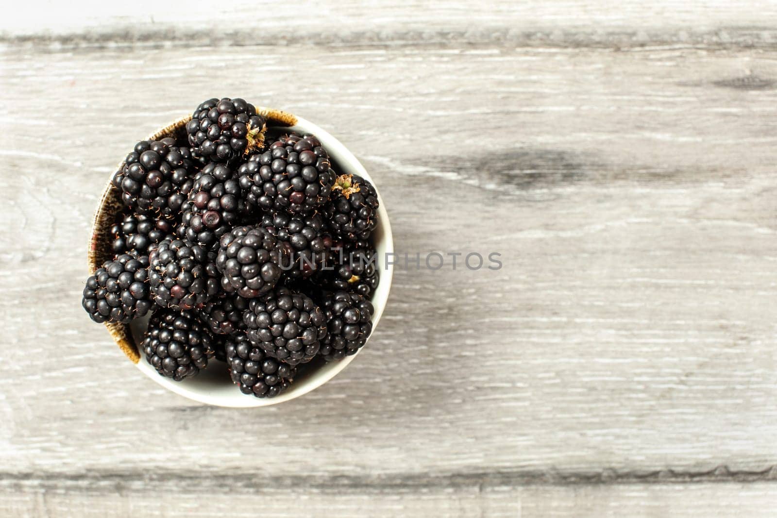 Tabletop view, small ceramic cup with blackberries on gray wood desk. Space for text on the right.