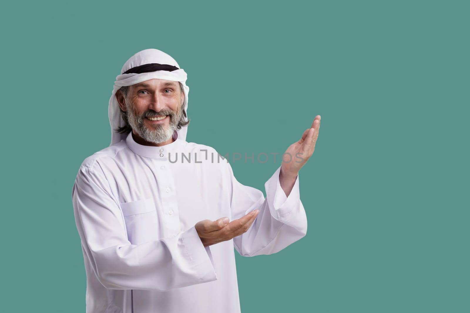 Joyful Arab man wearing Dishdasha, traditional clothing, smiles and points both palms towards copy space on green background. He is isolated and ready to add meaning to your project. by LipikStockMedia