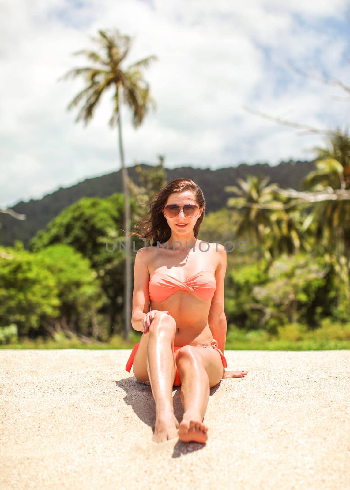 Young sporty woman in orange bikini and sunglasses sits on fine beach sand, wind in her hair, palm tree and jungle behind her.