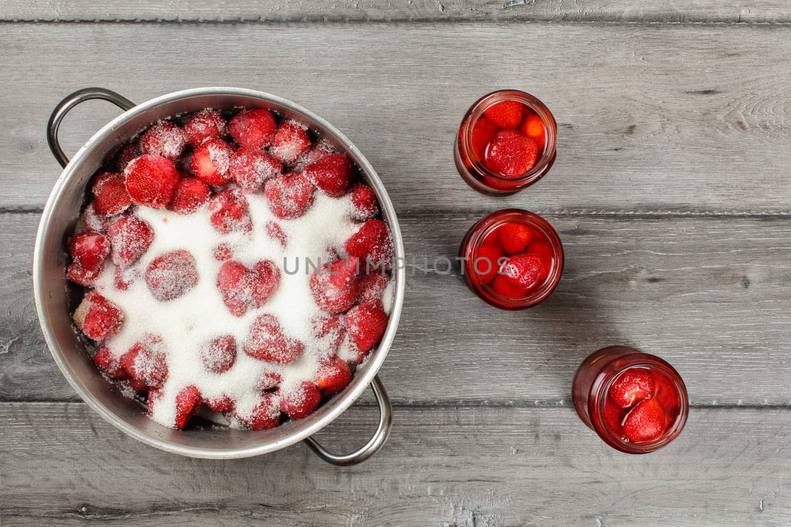 Tabletop view, steel pot with strawberries crystal sugar on top, three bottles with pickled fruit next to it. Homemade strawberry compote preparation.