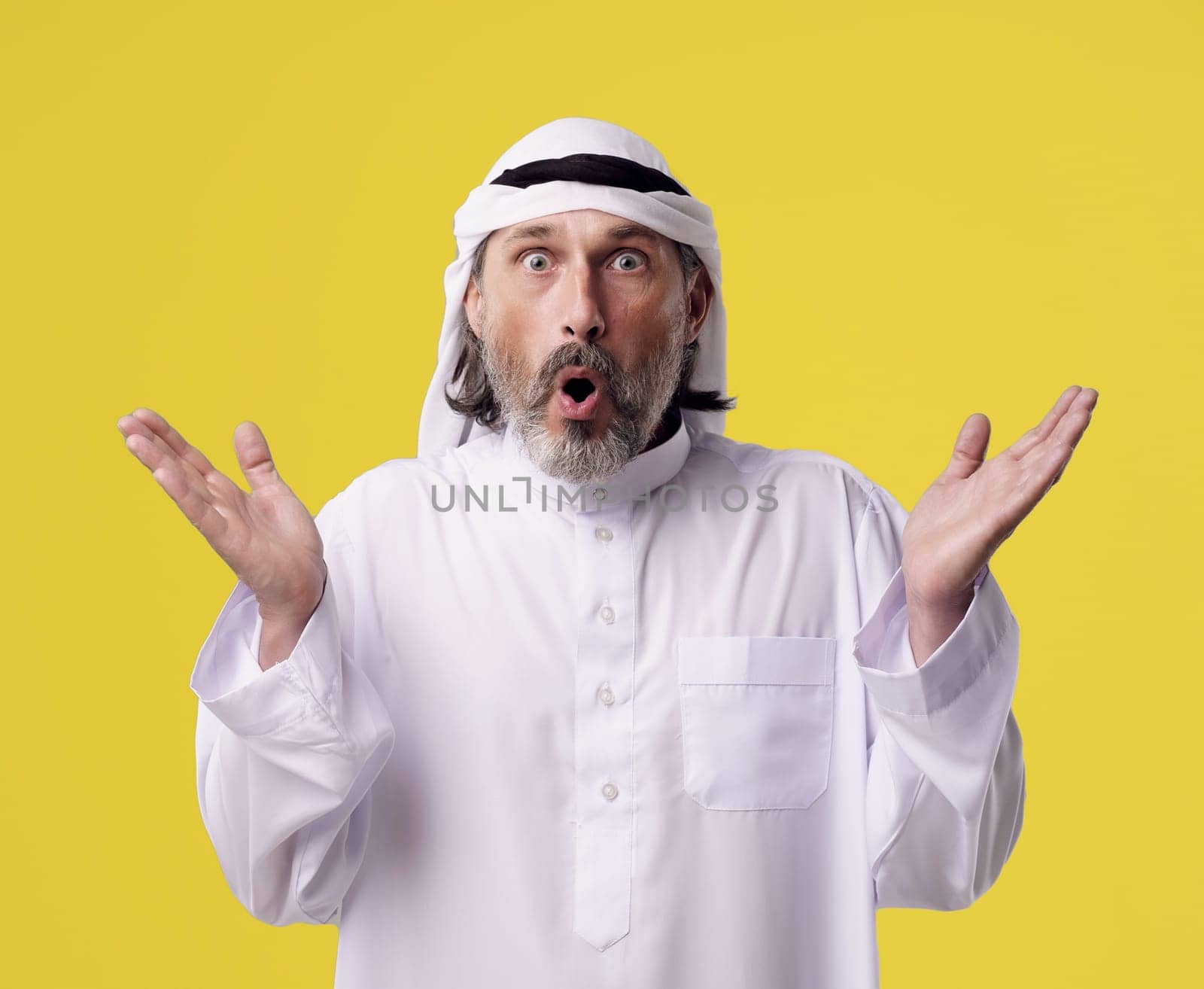 Surprised Arab man in traditional Dishdasha clothing raises both hands in the air, isolated on a bright yellow background with copy space. by LipikStockMedia