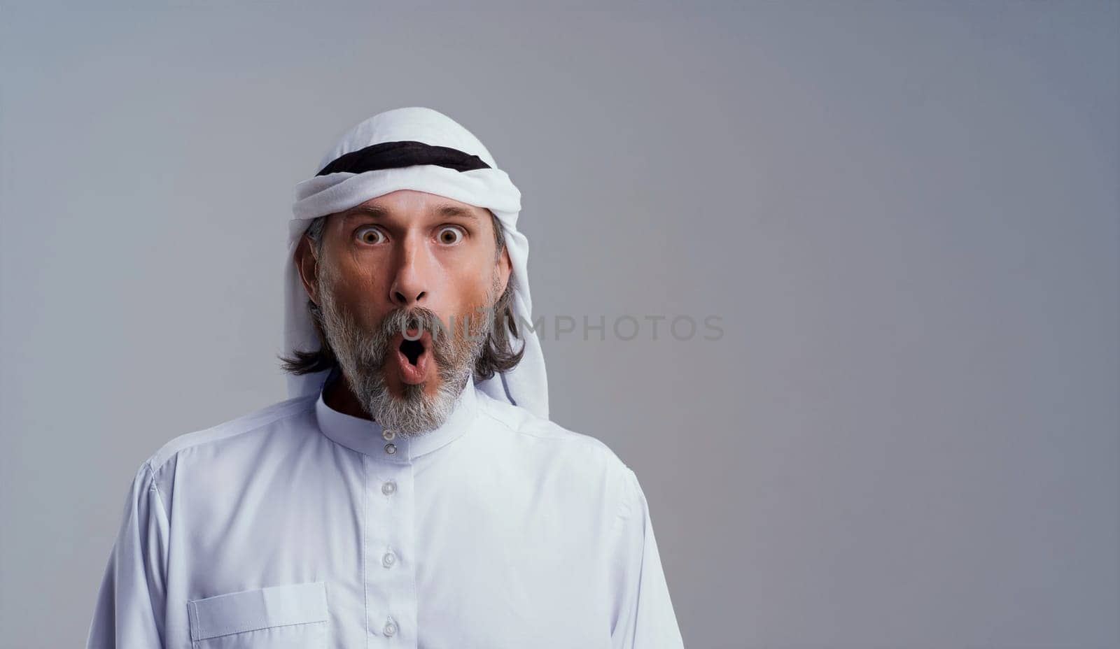 Surprised exiting Arab man wearing Dishdasha, traditional Islamic clothing, standing on toned blue background with copy space. High quality photo