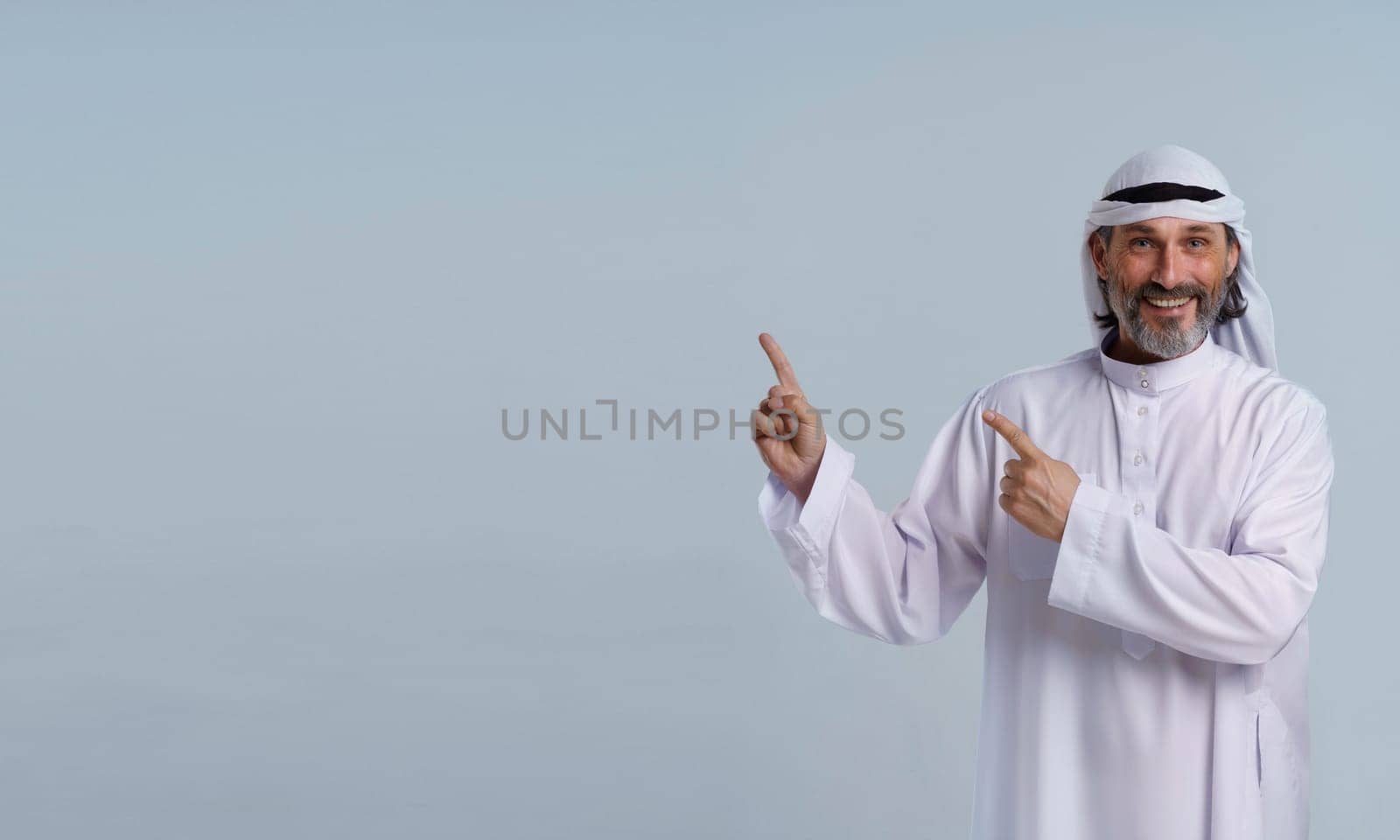 Arab man is with copy space. He is pointing his hands aside, indicating leadership, direction, and effective communication. Mid-length portrait isolated against blue background. by LipikStockMedia