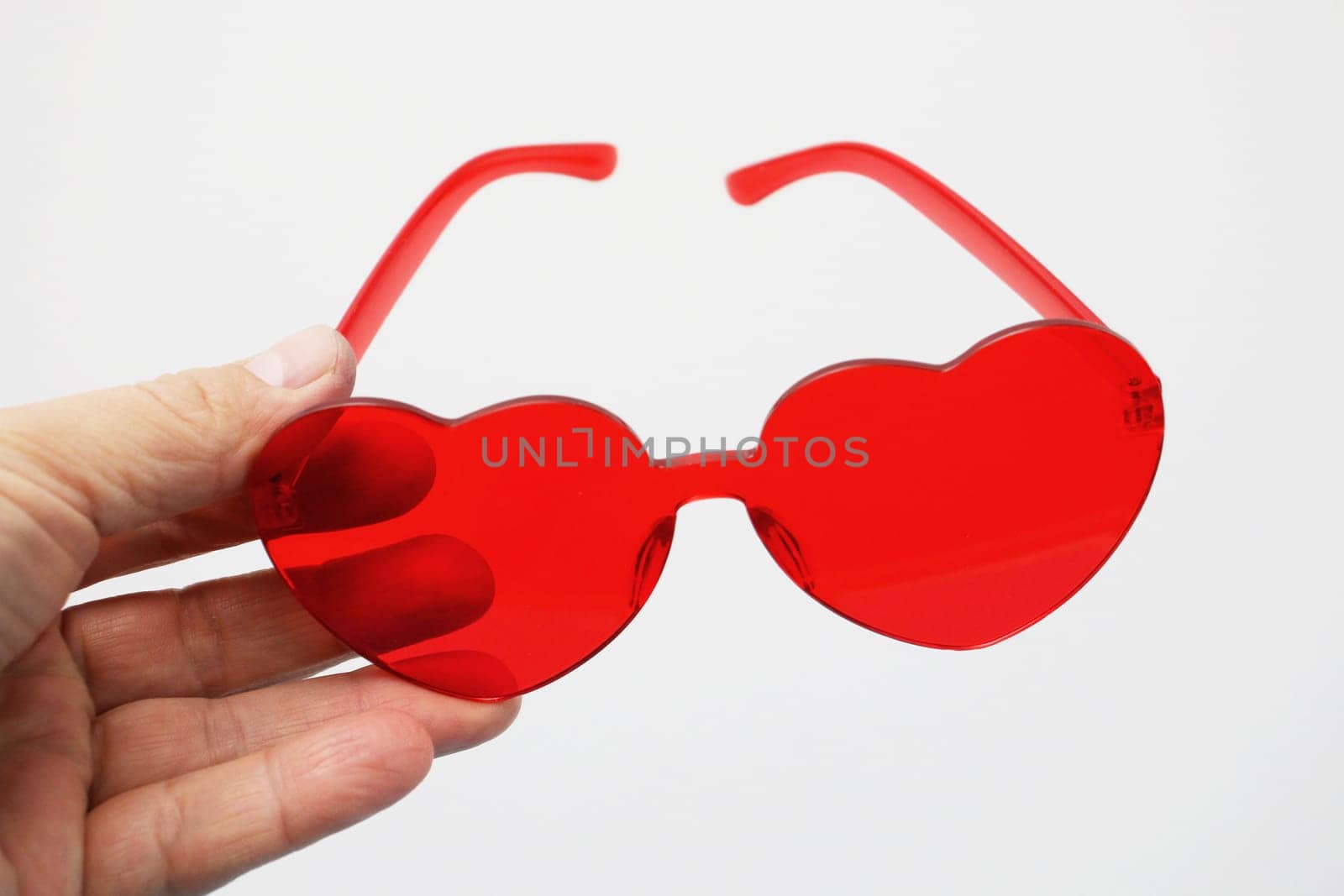 A hand holds red transparent sunglasses in the shape of a heart on a white background.