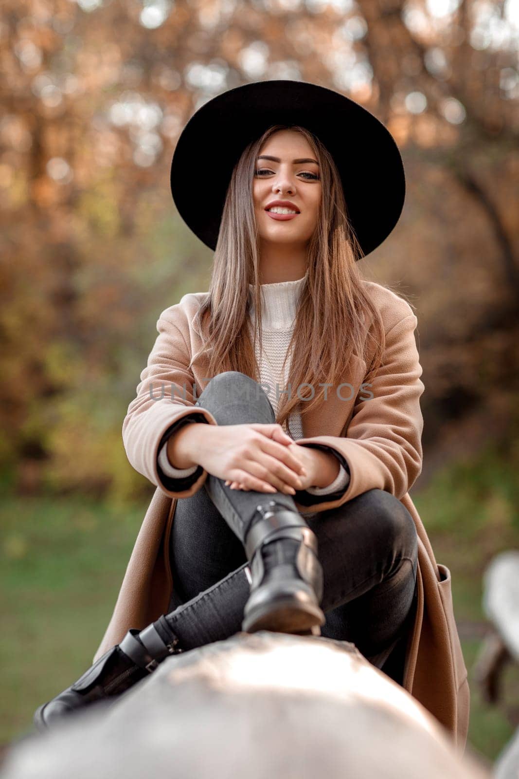 beautiful fashion woman in beige coat and black hat sitting on tree on sunny autumn day.