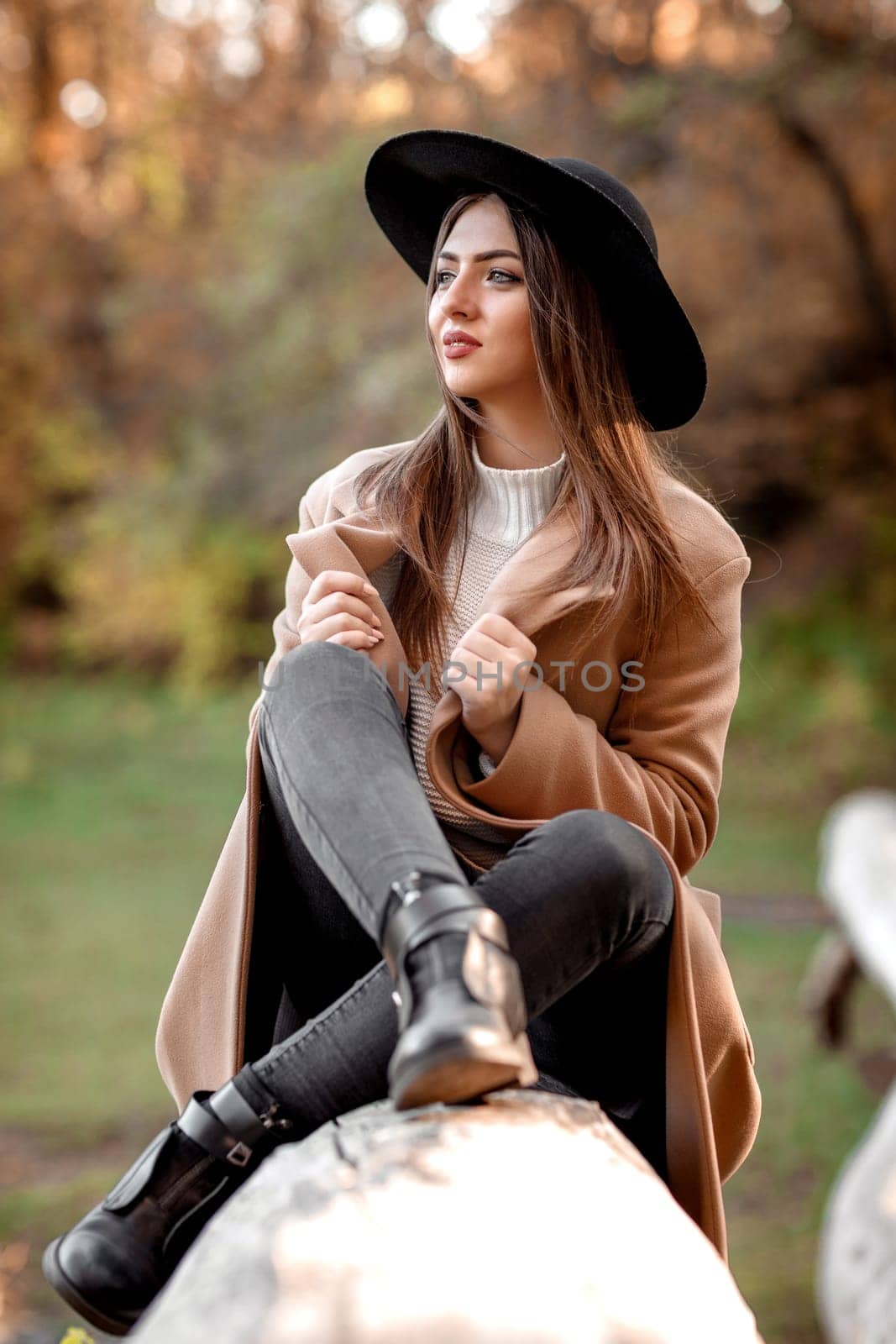 beautiful fashion woman in beige coat and black hat sitting on tree on sunny autumn day.